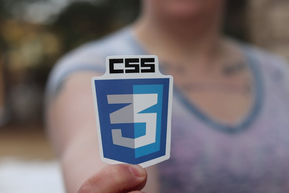 A person holding a sticker featuring the CSS3 logo, which displays a blue shield with the number 3 in white, and the acronym 'CSS' in bold black letters at the top. The individual is dressed in a casu