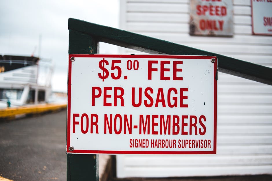 A close-up image of a white rectangular sign with bold red text indicating a $5.00 fee per usage for non-members, mounted on a green pole at a harbor or seaside location. In the background, there are