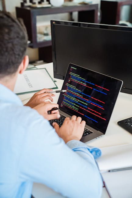 A person working indoors at a desk with two monitors and a laptop, actively engaged in computer programming. The laptop screen displays colorful lines of code, while the larger monitor beside it is tu